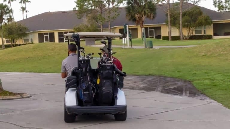 Golf cart with two people and golf bags on a paved path near a grassy hill and building.
