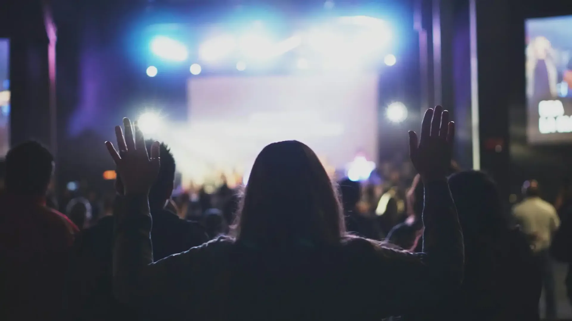 People with raised hands facing a brightly lit stage in a dark hall.