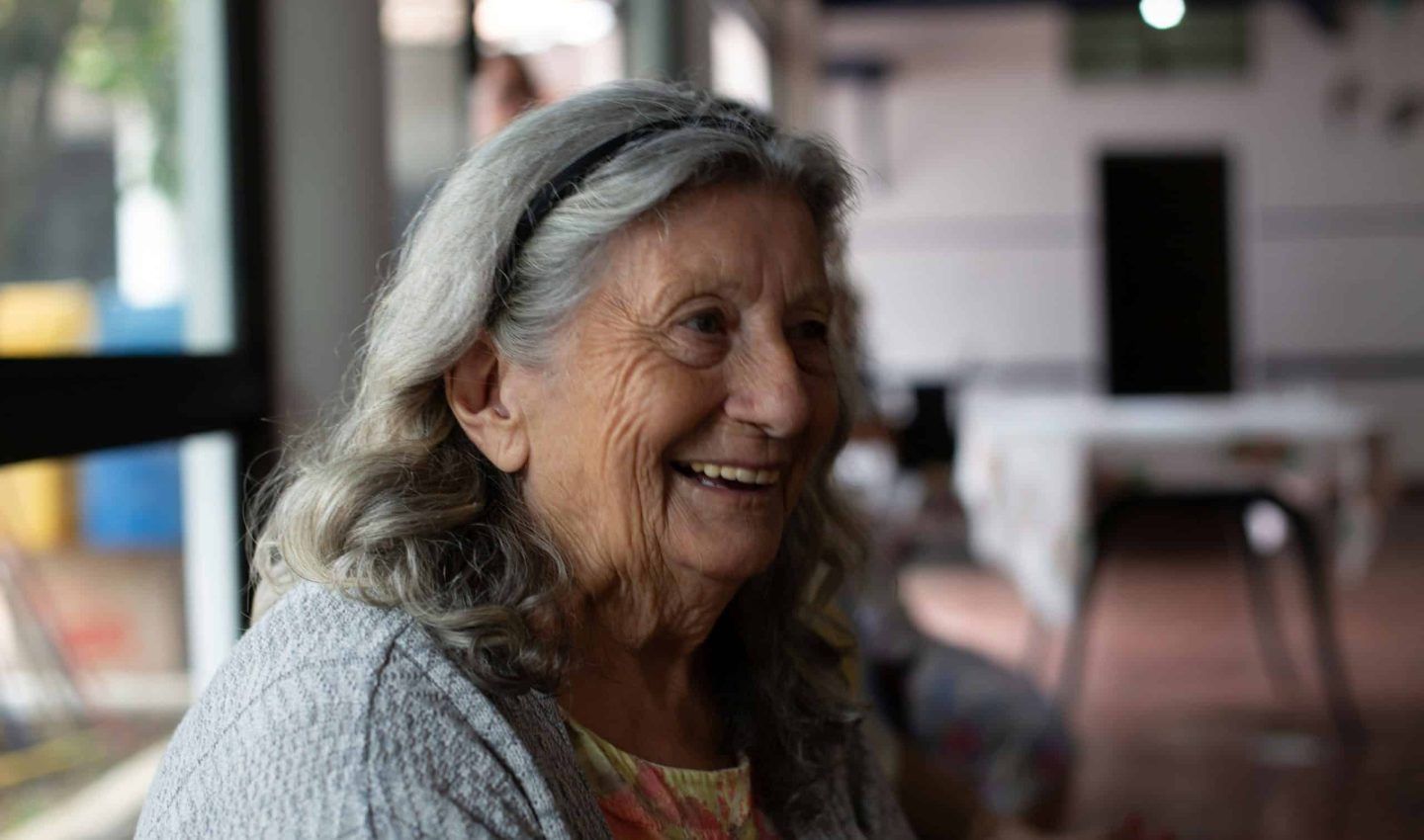Woman with gray hair and a headband smiles widely indoors.