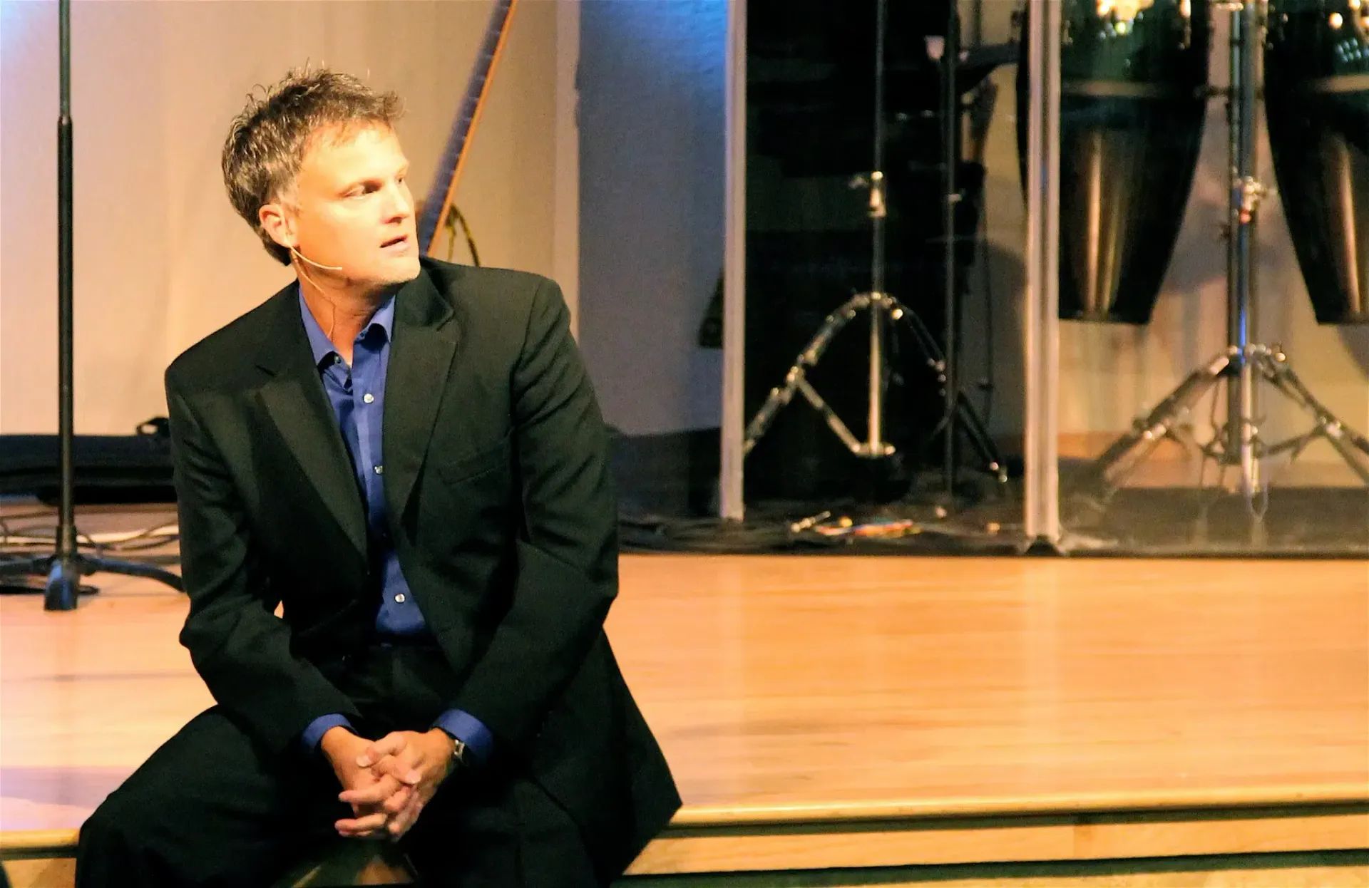 Man in suit seated on stage, looking upward. Background includes a drum set and lights.
