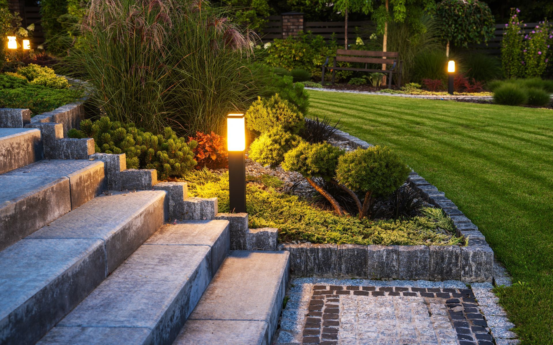 Stone steps lead into a garden at dusk, illuminated by glowing path lights beside a manicured lawn and landscaping.