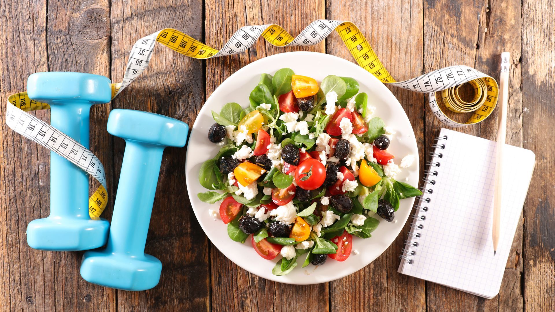 A plate of salad , dumbbells , measuring tape and a notebook on a wooden table.