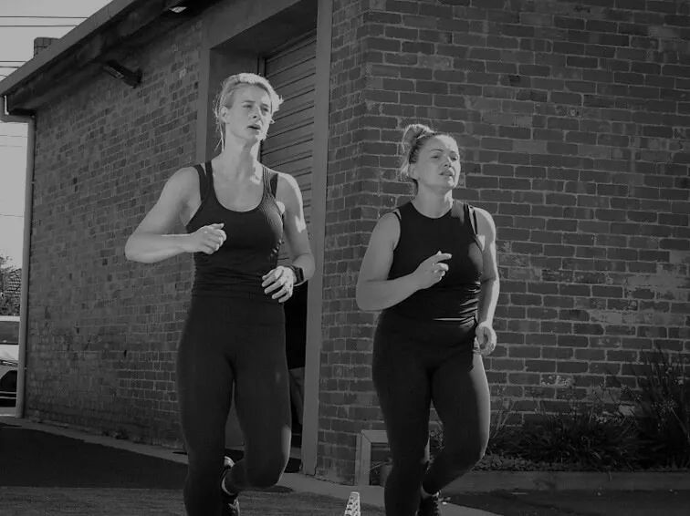 Two women are running in front of a brick building in a black and white photo.