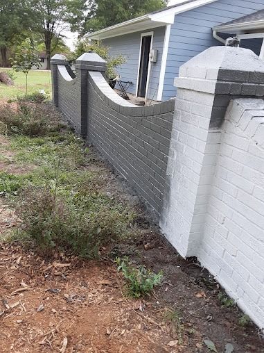 A grey brick wall curves along a yard next to a light blue house. Tall brick posts frame the wall.