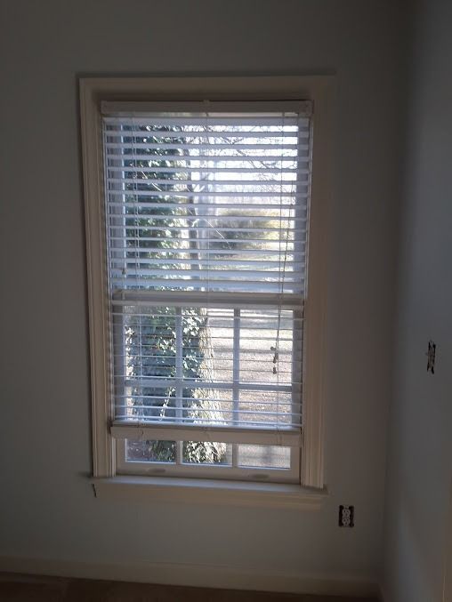 Window with white blinds, framed by a white trim, looking out onto a sunlit yard with trees.