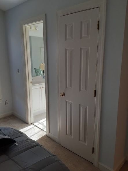 Bedroom with white door, bathroom door open, light blue walls, bed, and carpet.