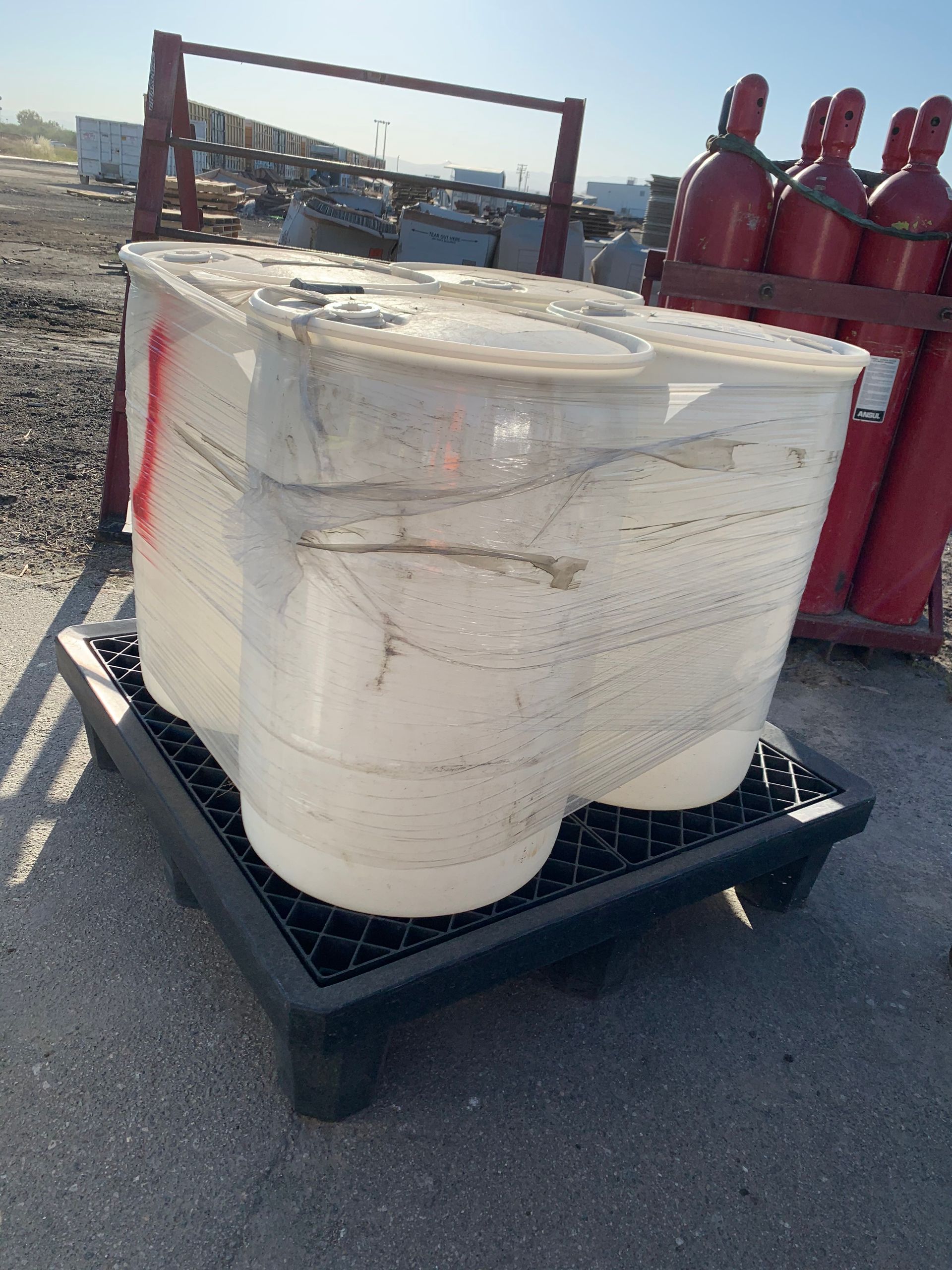 White container wrapped in plastic on a black pallet, with red cylinders in the background.