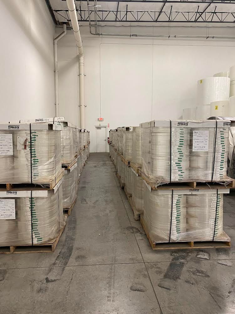 Warehouse with rows of palletized paper rolls, white walls, and a distant exit sign.