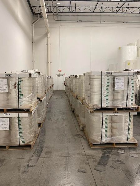 Warehouse with rows of palletized paper rolls, white walls, and a distant exit sign.