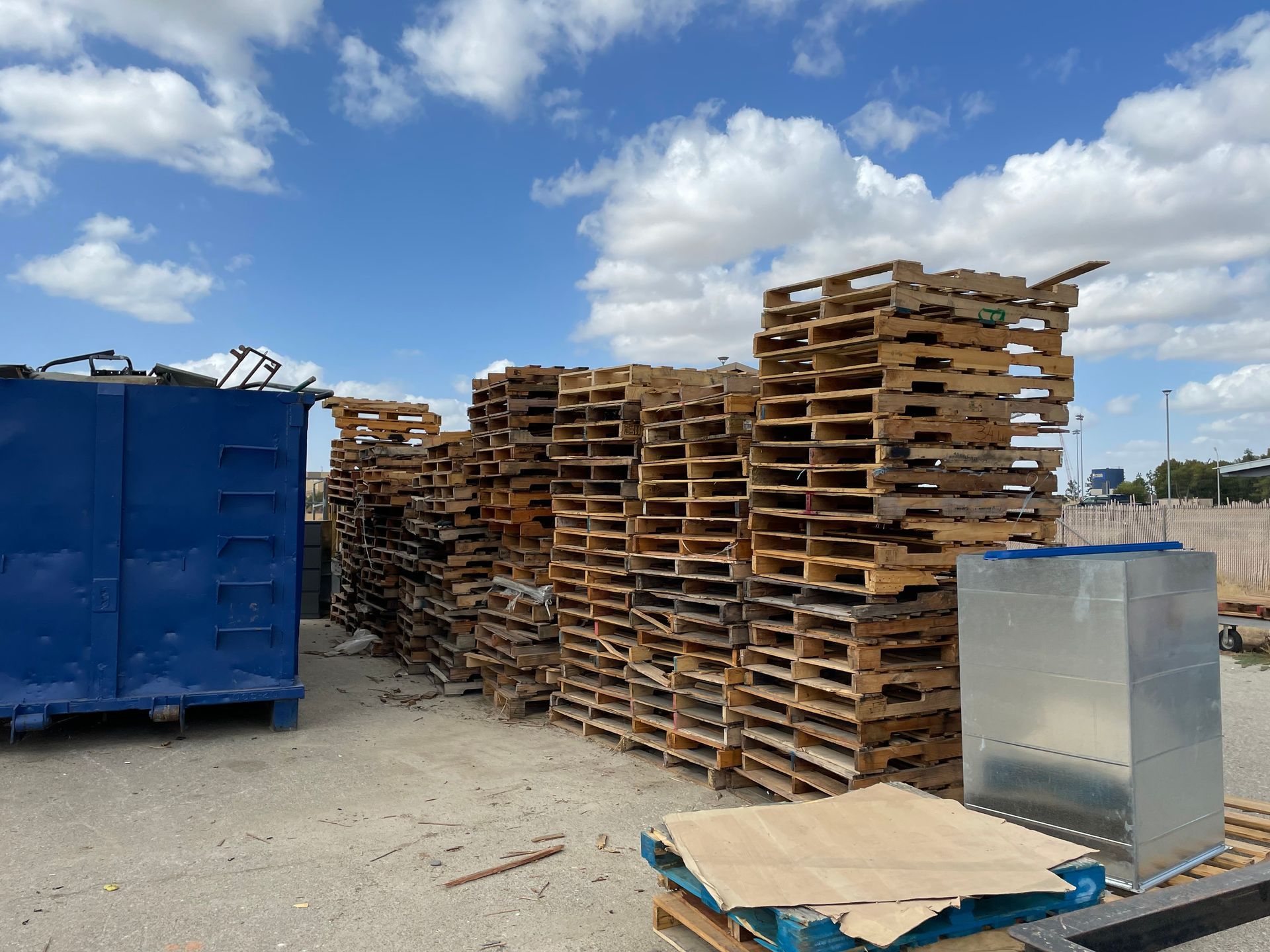 Stacks of wooden pallets outdoors, next to a blue dumpster and metal container, under a cloudy sky.