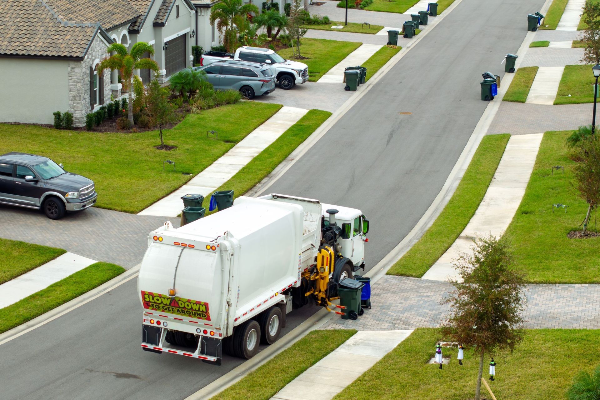 Garbage truck collecting trash bins along a suburban street. Green bins line the road with houses and cars.
