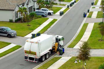 Garbage truck collecting trash bins along a suburban street. Green bins line the road with houses and cars.