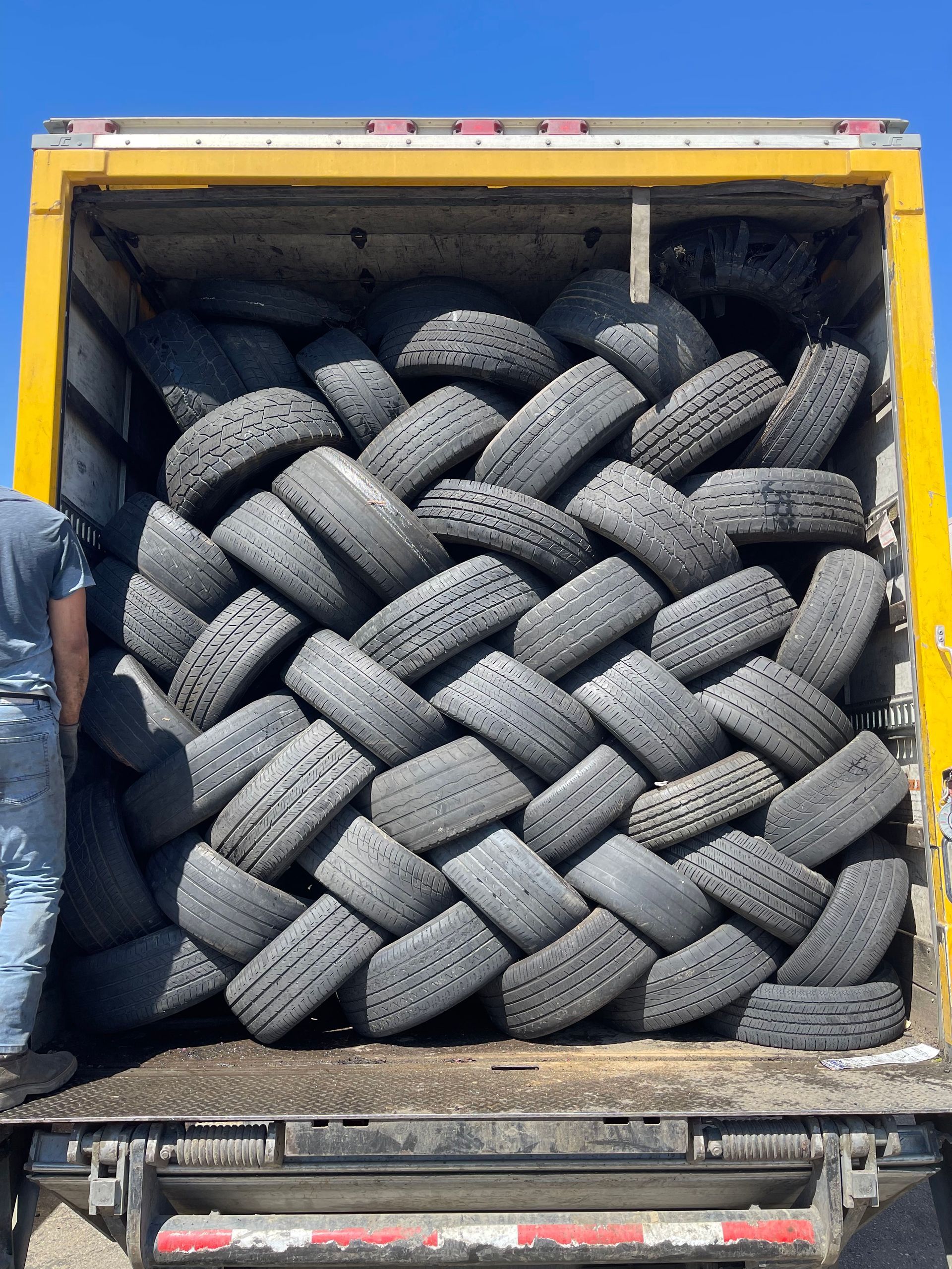 Used tires stacked in a truck bed, forming a tight, interlaced pattern. Someone in blue jeans and shirt is on the left.