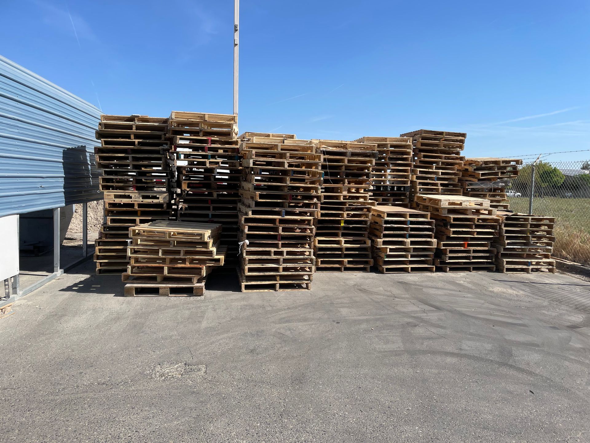 Pile of wooden pallets stacked outside against a blue sky, near a building.