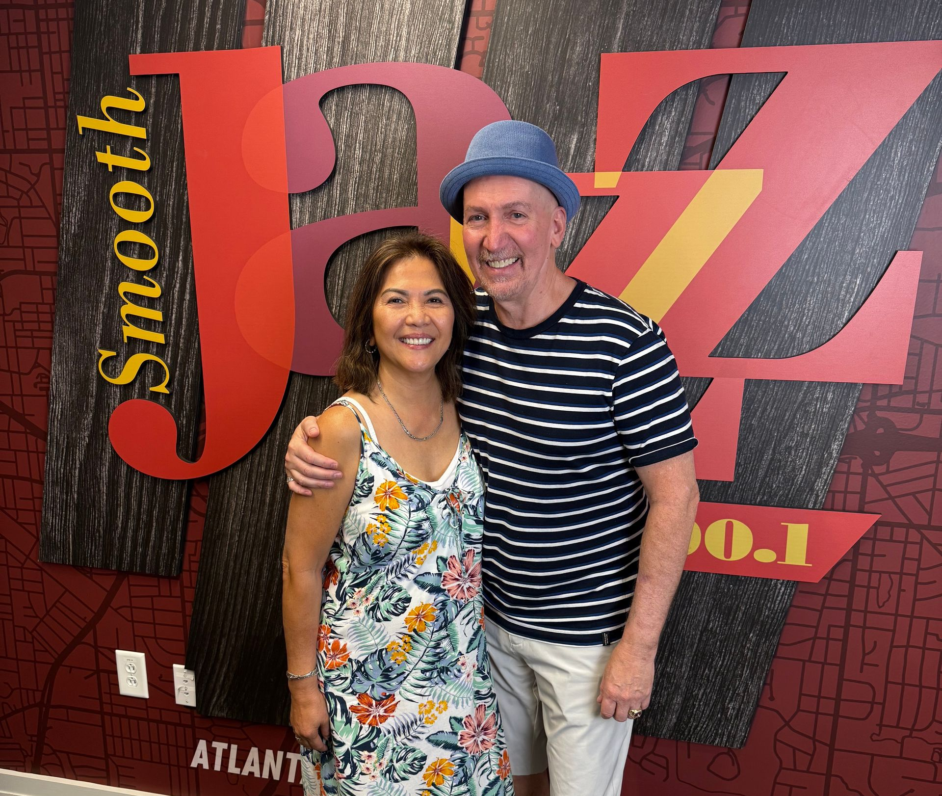 A man and a woman pose together in front of a Smooth Jazz 100.1 radio station backdrop in Atlanta.