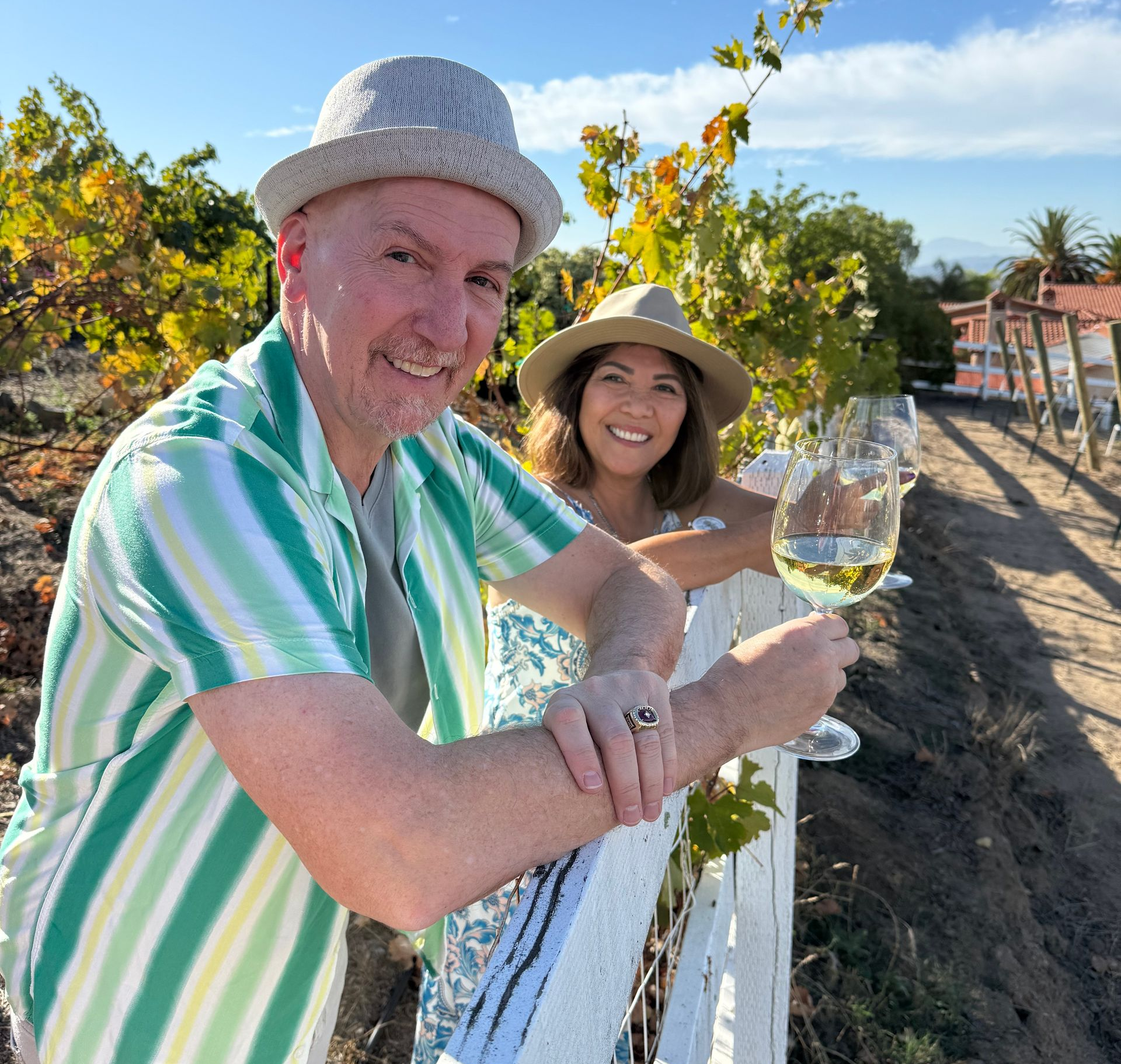 A person in a striped shirt and hat smiles while holding a glass of white wine at a vineyard with another person nearby.