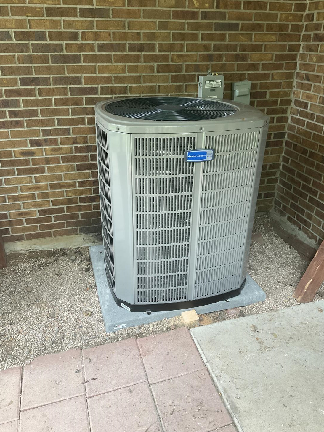 A gray air conditioner is sitting in front of a brick wall.