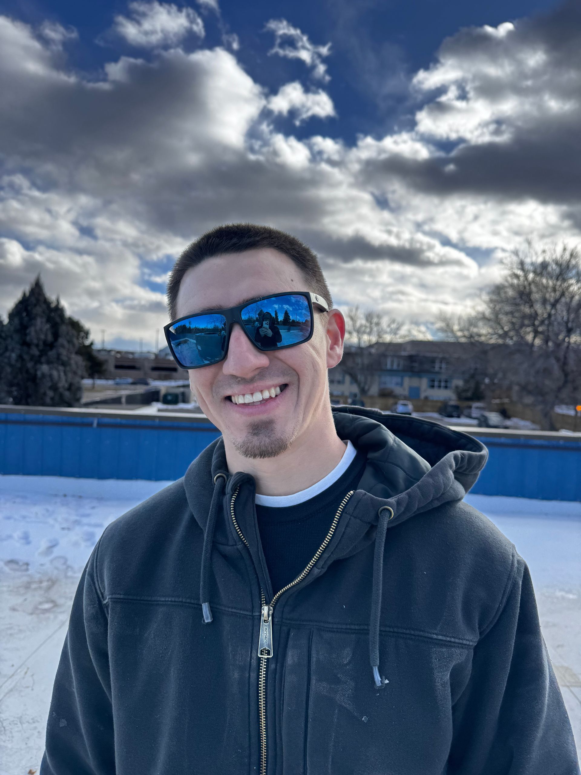 A man wearing sunglasses and a hoodie is smiling in the snow.