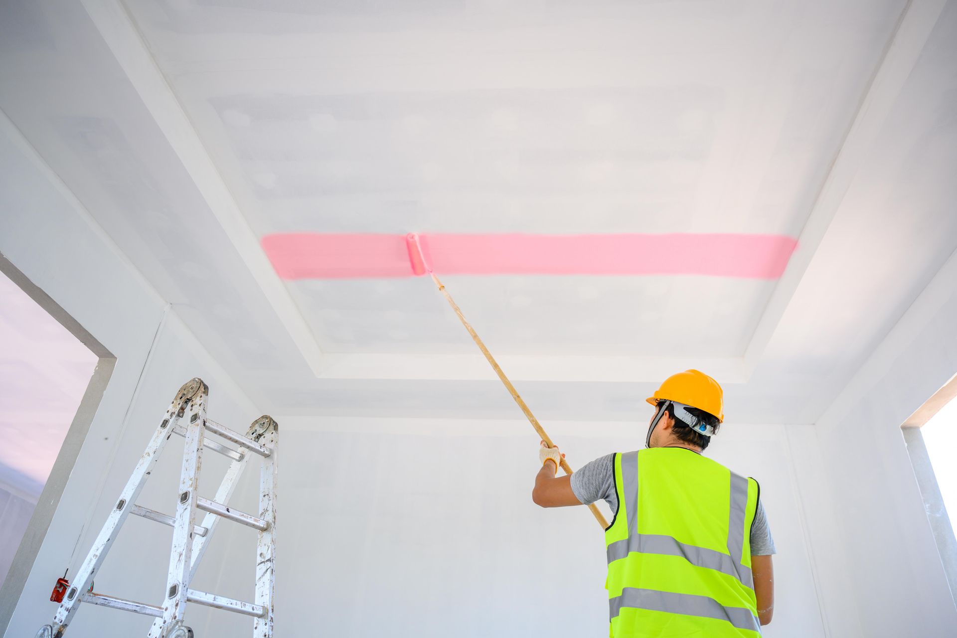Painter in a hard hat and safety vest using a roller to paint a pink stripe on a white ceiling.