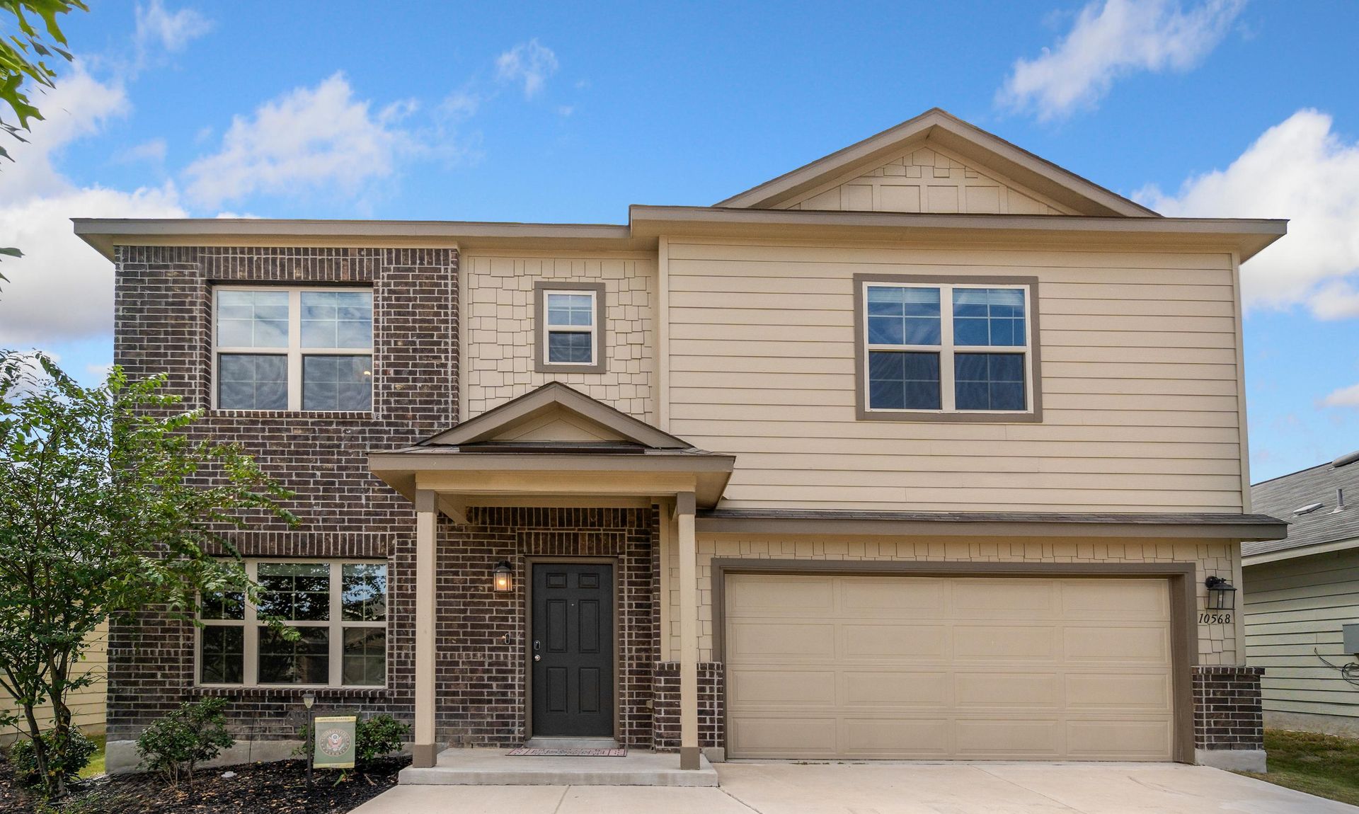 Two-story beige and brick house with a garage and front porch, set against a blue sky.