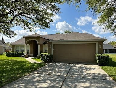 Tan house with a brown garage door, surrounded by green lawn and trees on a sunny day.