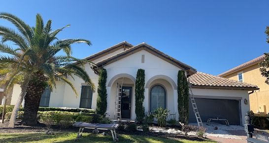 House with light-colored stucco exterior, brown roof tiles, and a palm tree against a bright blue sky.