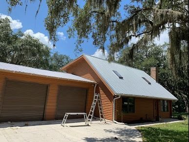 A brown house with a metal roof, two garage doors, and a ladder on the side, under a tree on a sunny day.