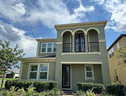Two-story house with olive green exterior, balcony, and dark door under a cloudy sky.