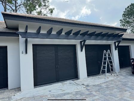Garage with black doors, trim, and pergola against white walls. A ladder leans against the middle door.