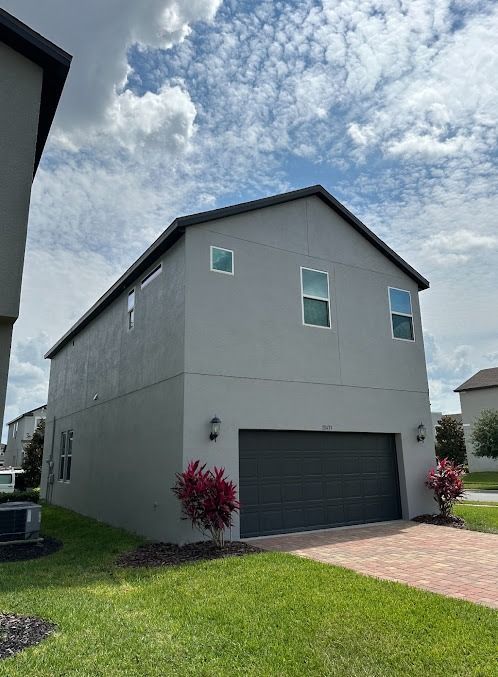 Gray two-story house with a dark gray garage door, brick driveway, and small bushes. Sky above.