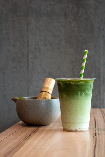A cup of matcha latte with a straw and a bowl of green tea on a wooden table.