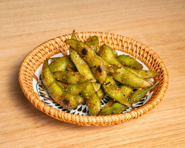 A plate of edamame beans in a wicker basket on a wooden table.