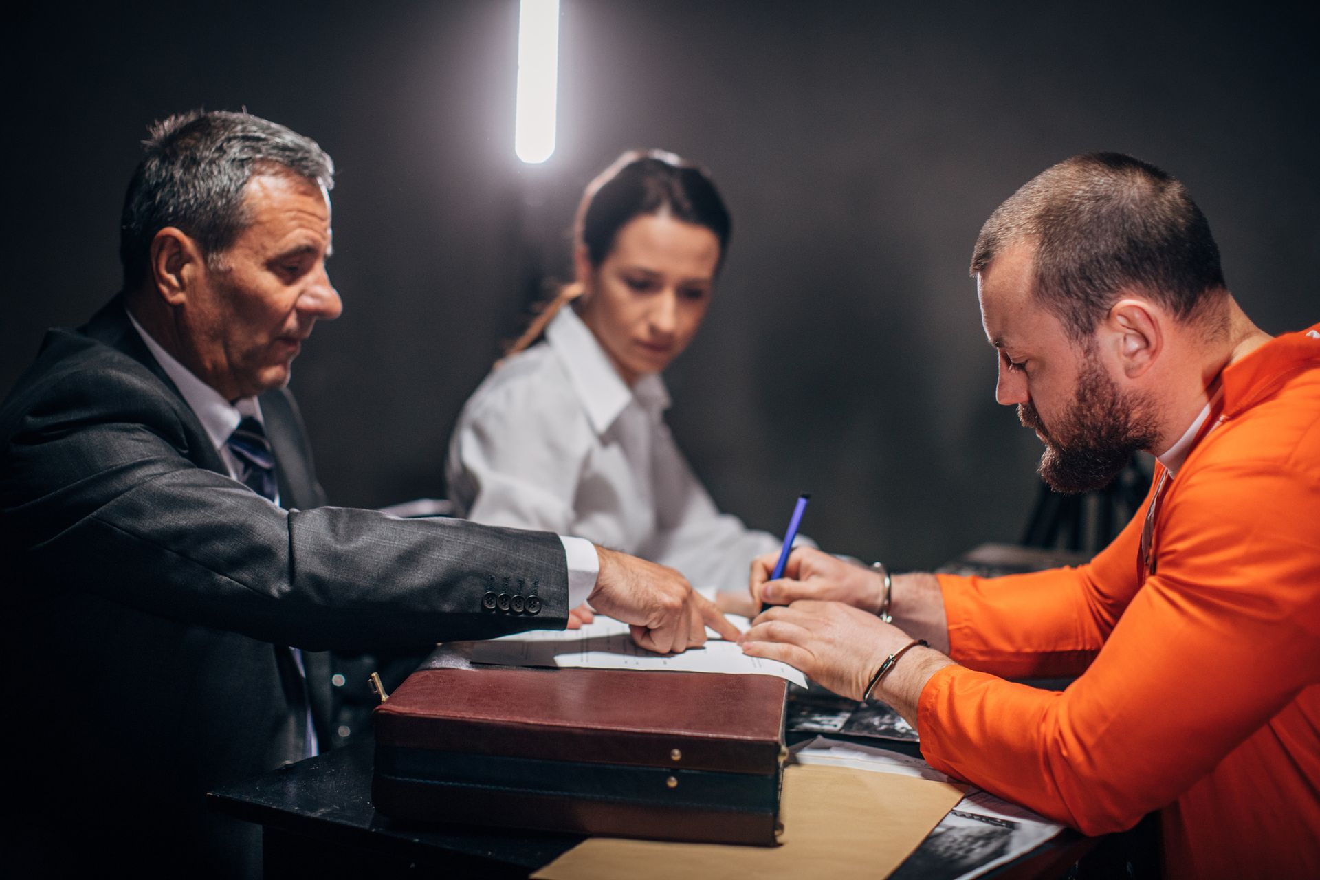 A man in an orange prison uniform is sitting at a table with a lawyer and a woman.