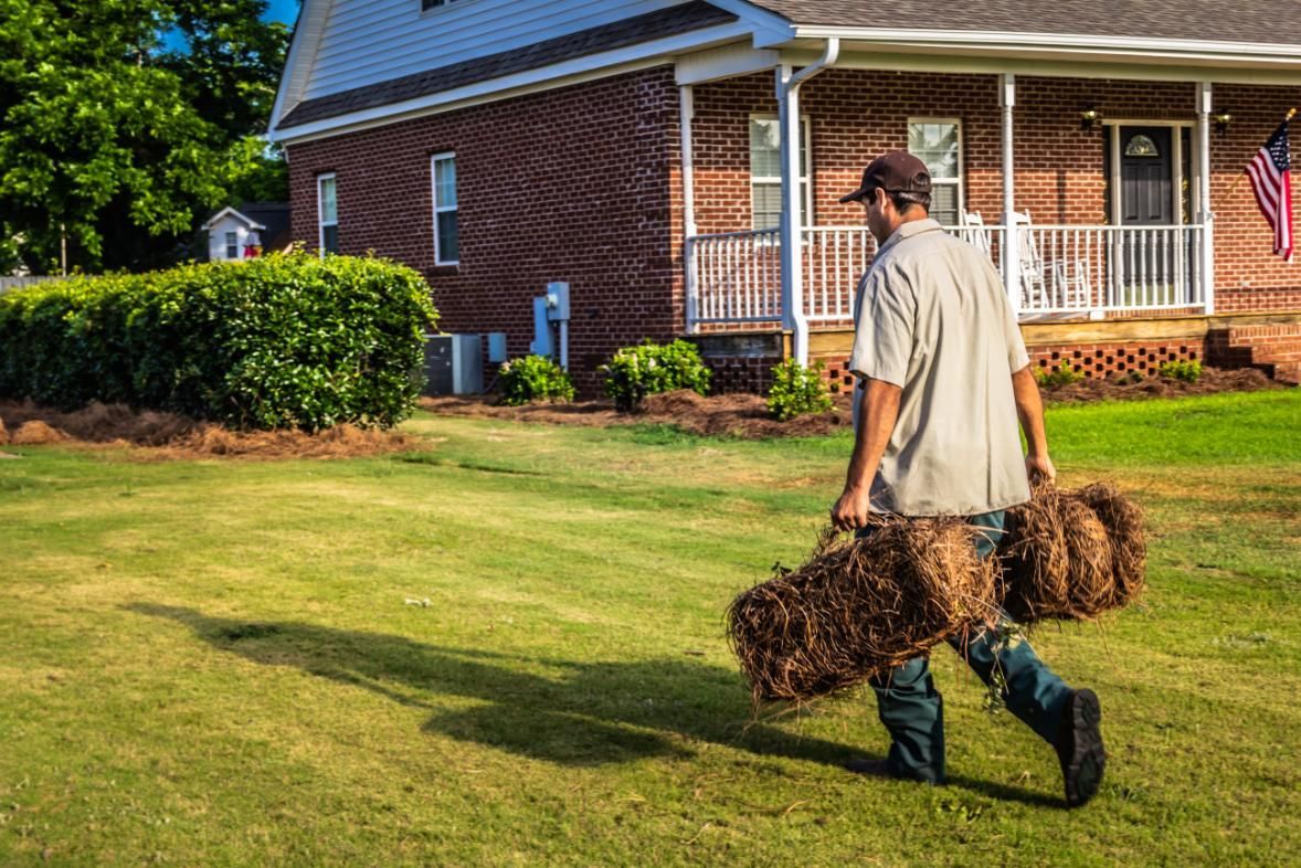 Man carrying two bundles of straw across a green lawn toward a brick house with a porch and American flag.