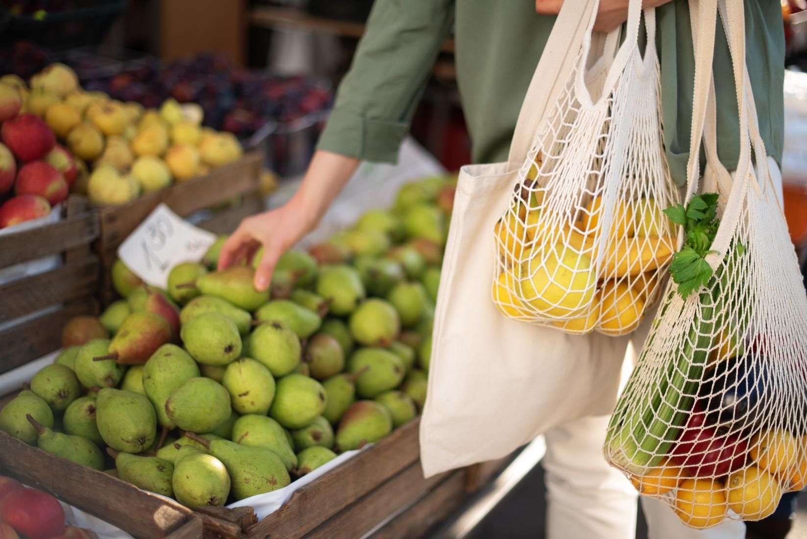 Person shopping at a market, selecting a pear and holding string bags filled with produce.