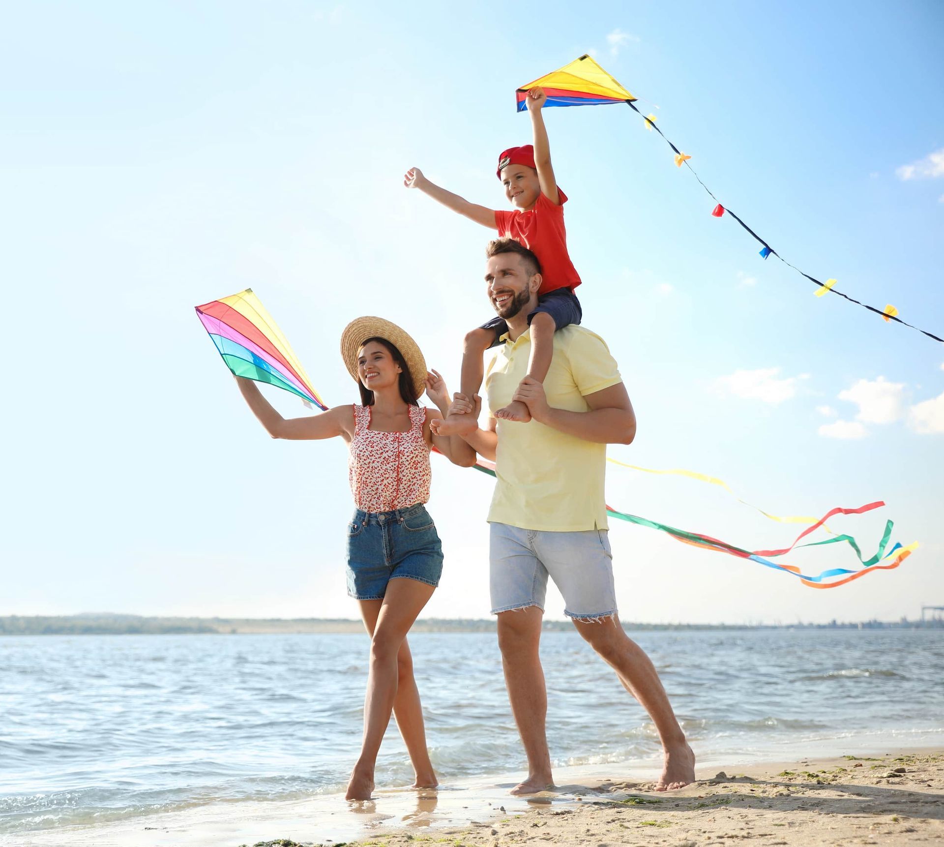 Family on beach flying kites, sunny day. Man carries child on shoulders. Woman holds kite.