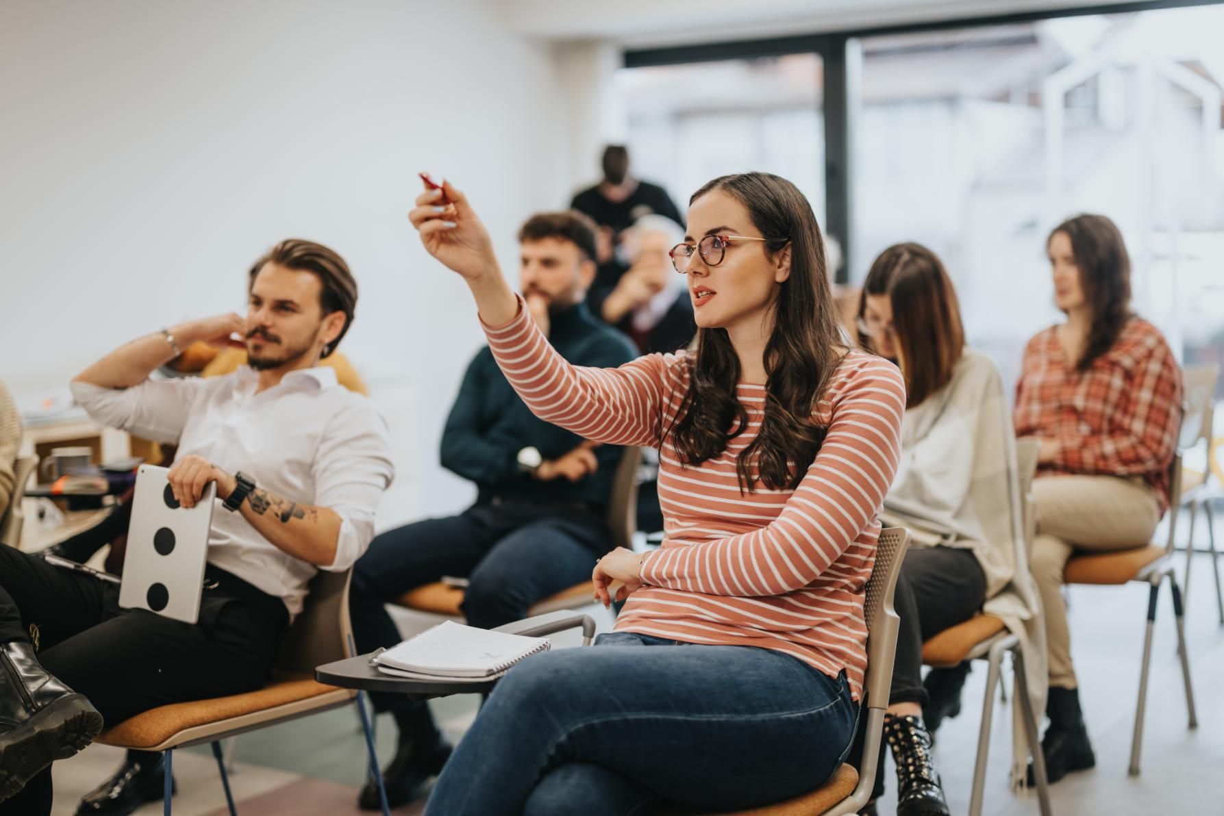 Woman in a striped shirt raises hand with pen in a classroom setting, other people sit and listen.