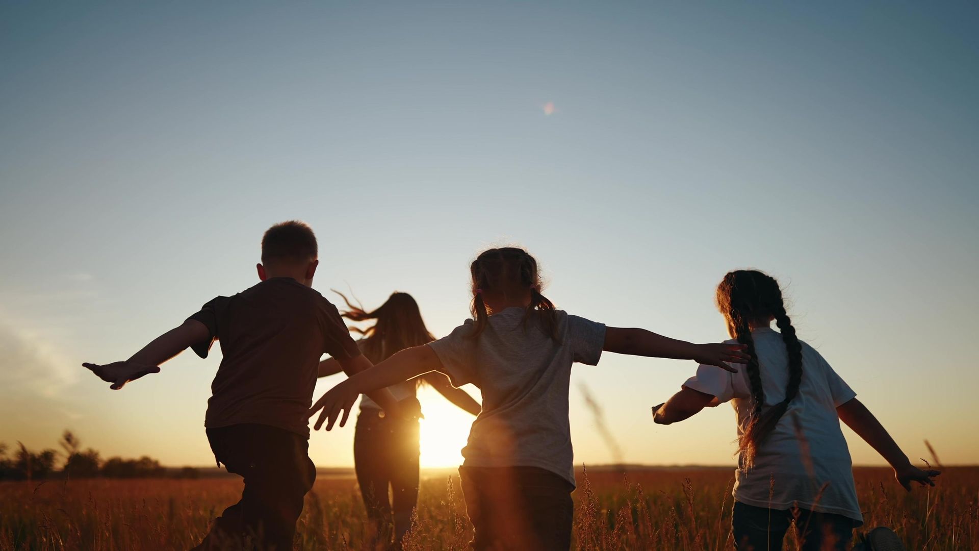 Children running with arms outstretched in a field at sunset, silhouetted against the sky.