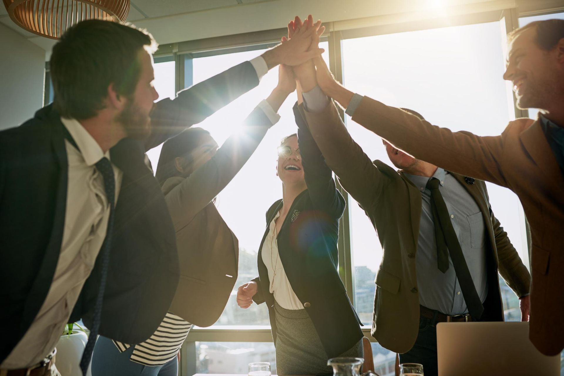 Business team celebrates success with a group high-five in a sunlit office.