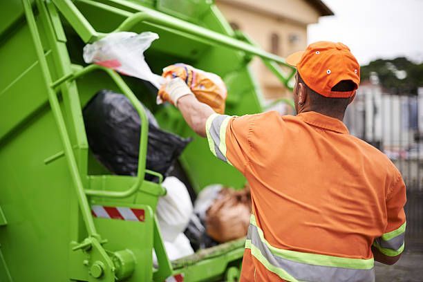 Garbage collector in orange uniform emptying a trash bag into a green recycling truck