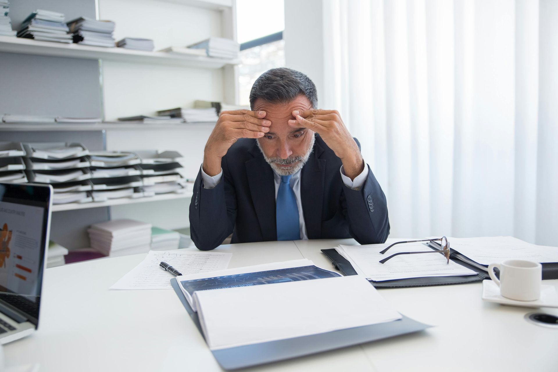 A man in a suit and tie is sitting at a desk with his hands on his head.