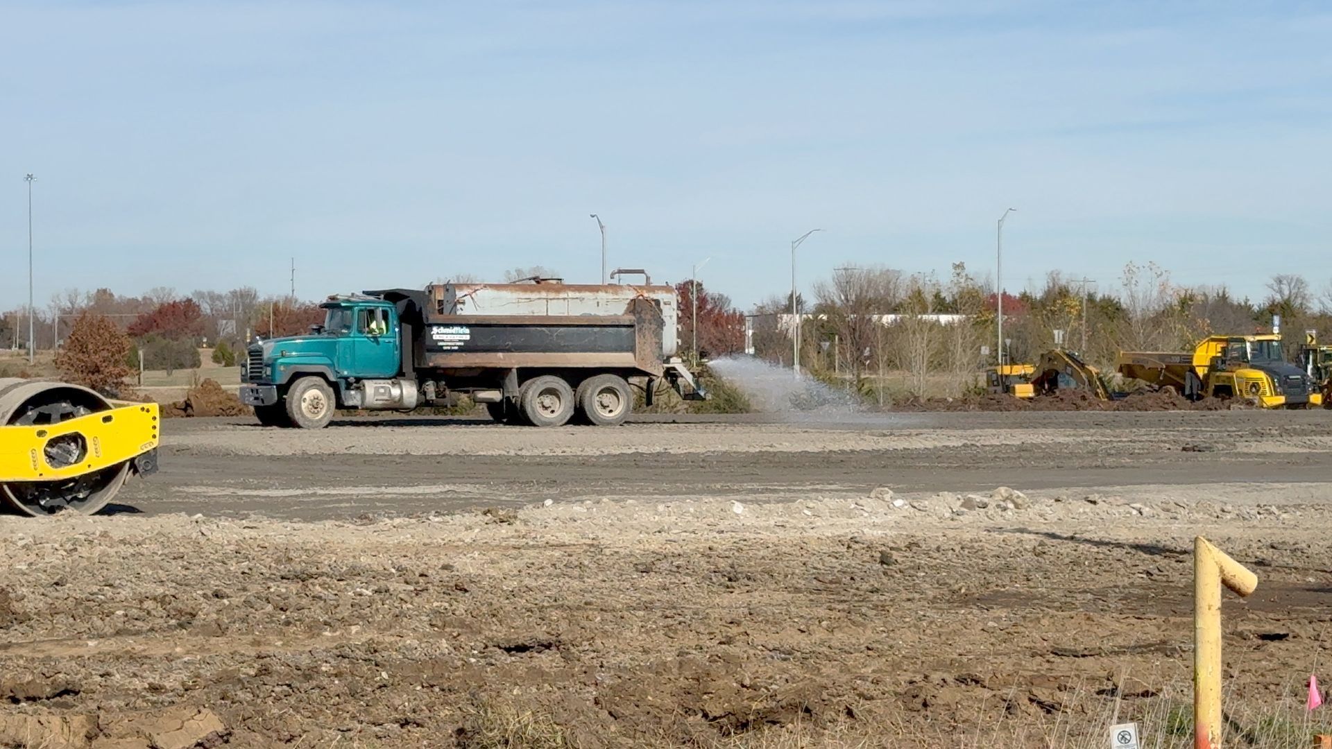 A truck is driving down a dirt road next to a yellow roller.