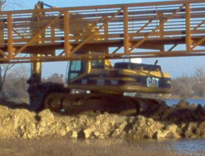 A yellow cat excavator is under a bridge over a body of water