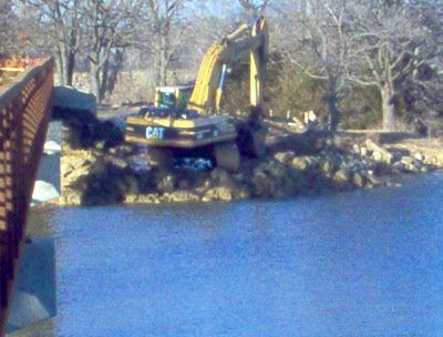 A cat excavator is working on a rocky shoreline near a body of water.