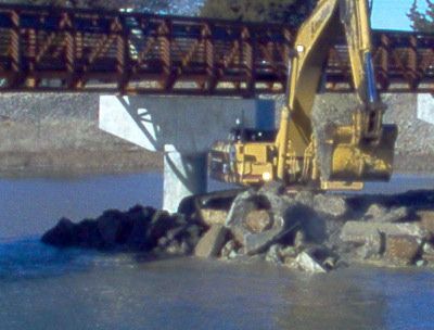 A yellow excavator is working on a bridge over a body of water