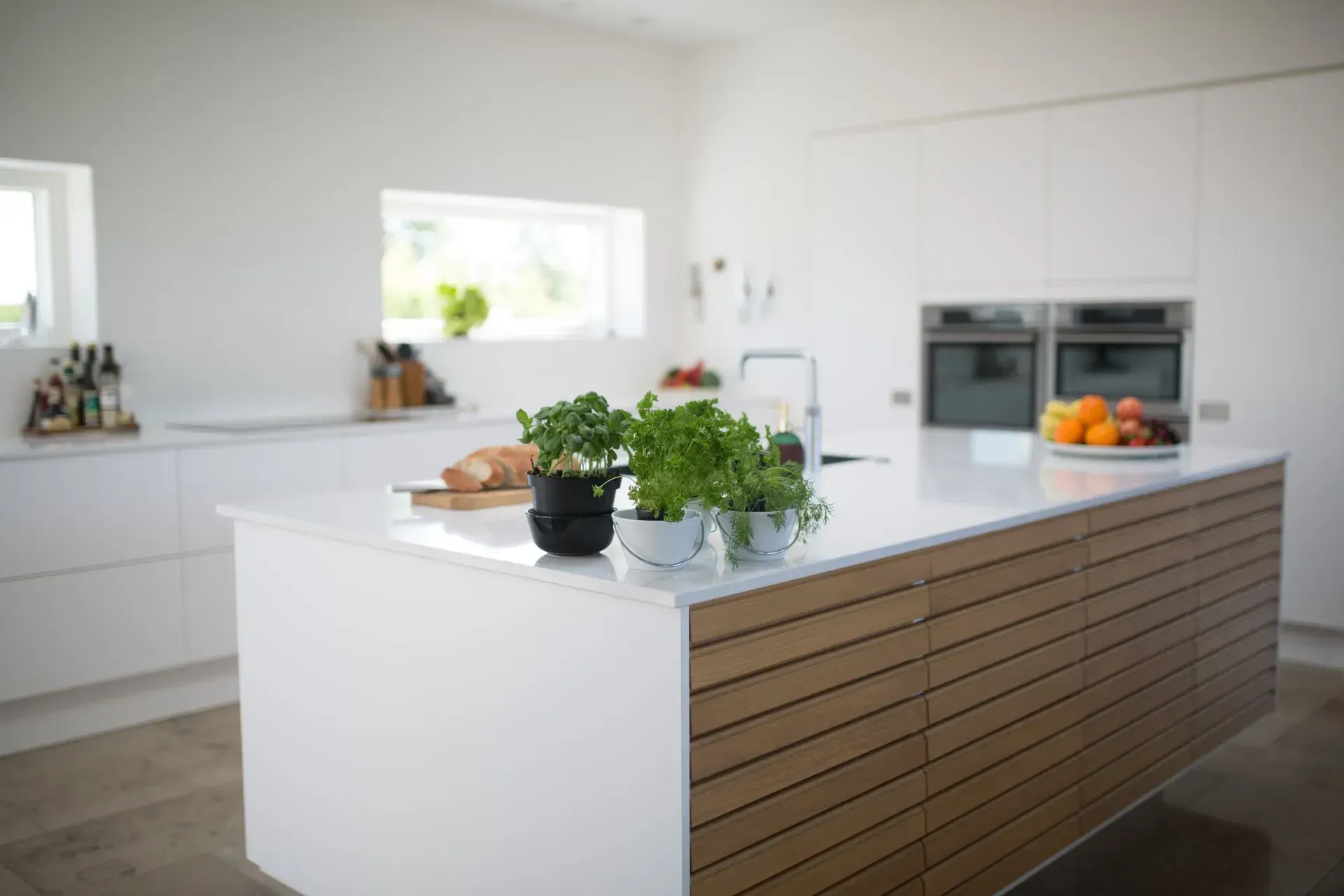 Modern white kitchen with a large island, built-in oven, and fruit and herbs on the counter