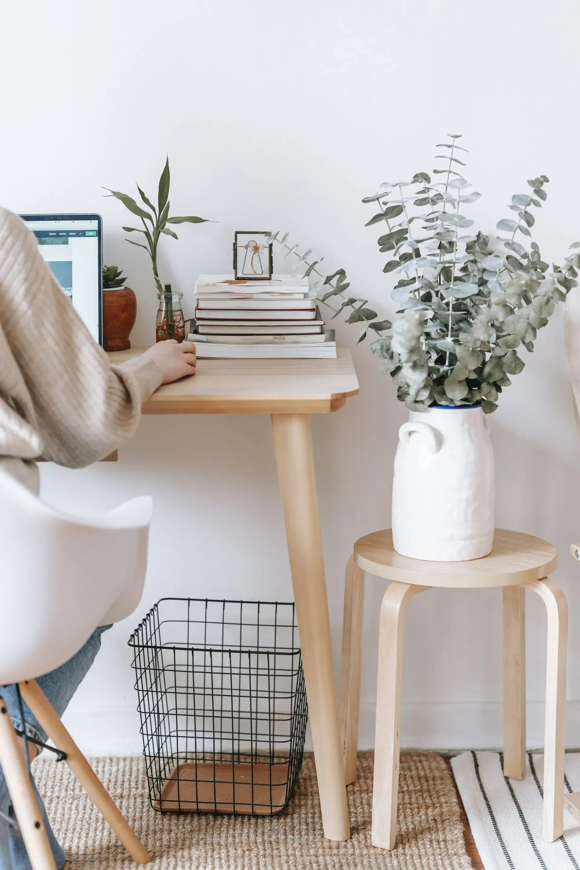 Minimal desk setup with laptop, stacked books, plants, and a white vase in a bright room