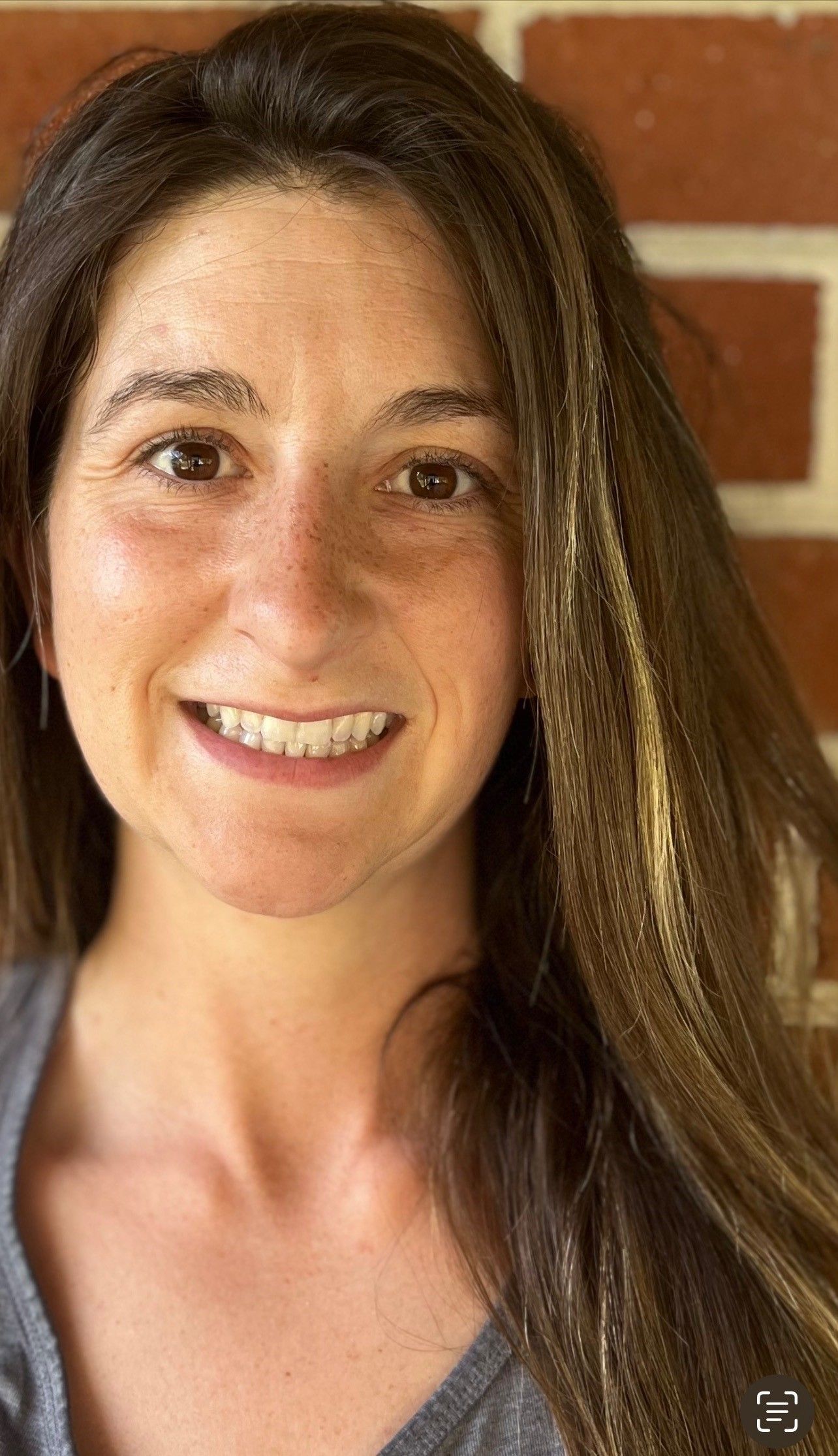 A woman with long hair is smiling in front of a brick wall.