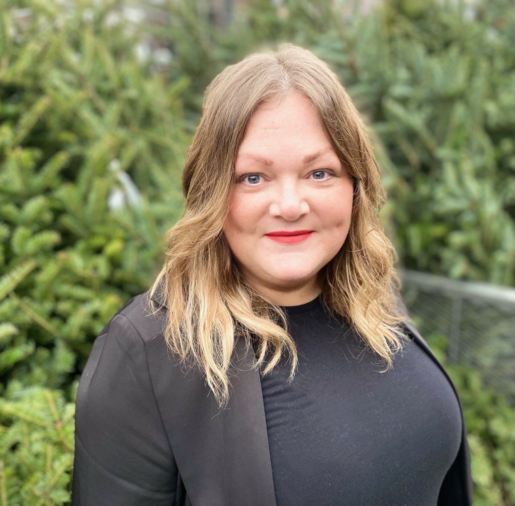 A woman wearing a black shirt and a black jacket is standing in front of trees.