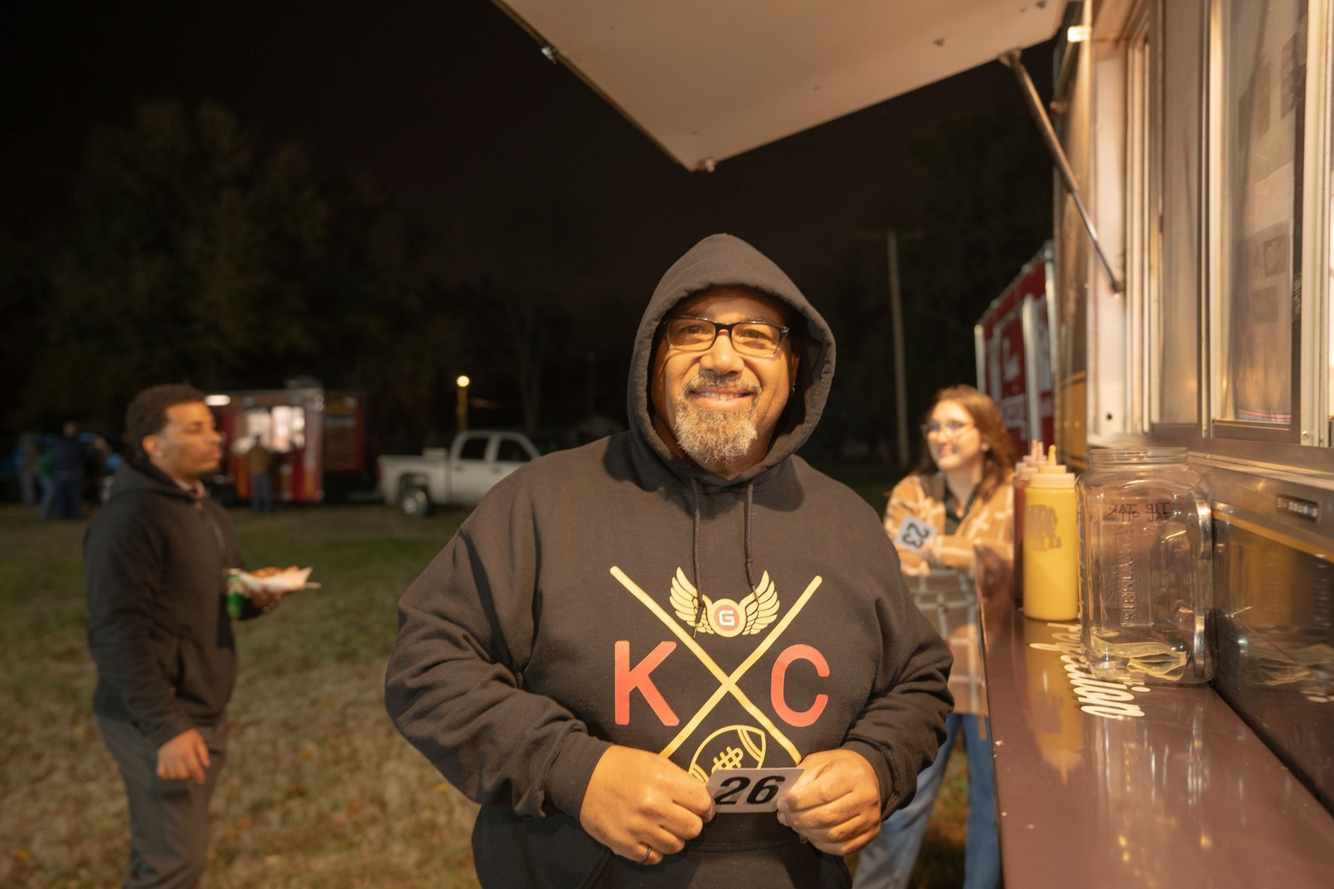 A man in a hoodie is standing in front of a food truck at night.